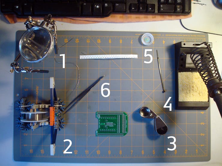 Going counter-clockwise from the upper left. 1: Third hand for holding parts in place. 2: Solder dispenser, made from K'Nex for geek-cred. 3: Jeweler's loupe for close inspection. 4: WES51 Solder Station. 5: A puddle of no-clean flux in a bottlecap with a little plastic stick to move it around. 6: Fine tipped tweezers.